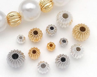 Puede incluir: Un surtido de cuentas de joyería en tonos dorados y plateados. Las cuentas tienen un diseño acanalado en forma de flor. Algunas cuentas están dispersas, mientras que otras están ensartadas con cuentas grandes, blancas y doradas, lo que sugiere un collar o pulsera.