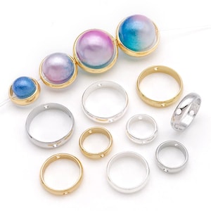 May include: An assortment of jewelry components, including gold and silver rings, and iridescent pearl-like beads set in gold-tone bezels. The beads display shades of blue, pink, and purple. The components are arranged on a white surface.