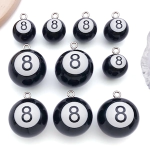 May include: A collection of black and white eight ball charms with silver-colored loops for jewelry making. The charms are round with a glossy finish, featuring the number "8" in white. The charms vary slightly in size.