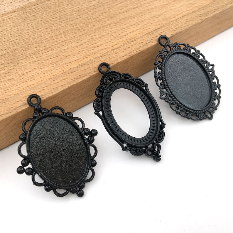 May include: Three black metal oval-shaped pendant trays with ornate designs. The trays have a smooth, black finish and are ready to be filled with resin, glass, or other materials.
