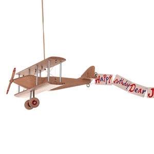 Puede incluir: Un avión de juguete de madera con una pancarta que dice "Happy birthday Dear JOHN". El avión de juguete tiene una hélice, ruedas y una cuerda para colgarlo. La pancarta tiene letras rojas y azules.