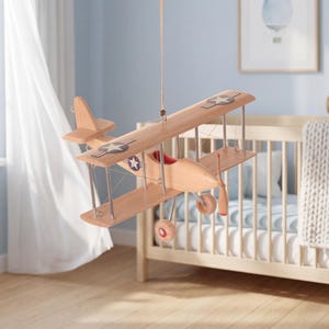 Peut inclure: Un avion jouet en bois avec des détails rouges et des décalcomanies d'étoiles est suspendu à une ficelle. L'avion jouet est suspendu devant une chambre d'enfant bleu clair avec un berceau et une fenêtre. C'est un jouet classique.
