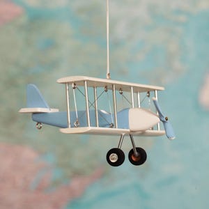 May include: A hanging biplane ornament featuring a light blue and white colour scheme. The model aircraft has two stacked wings, a front-facing propeller, and two black wheels attached to thin metal struts, suspended by a thin white string.