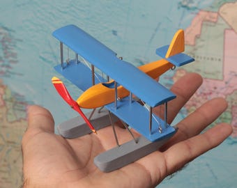 Könnte beinhalten: Ein kleines hölzernes Doppeldecker-Modell mit einem leuchtend orangefarbenen Rumpf, blauen Flügeln, einem roten Propeller mit gelben Spitzen und grauen Schwimmern. Das Flugzeug hat dünne Metallstreben und Drahtverspannungen.