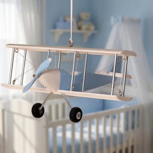 Puede incluir: Móvil de avión biplano de madera en un cuarto de bebé. El avión de juguete tiene una hélice y detalles en azul claro, con ruedas negras. El móvil está suspendido del techo, creando un elemento decorativo para la habitación del bebé. La envergadura es de unos 40 cm.