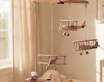 May include: A hanging nursery mobile featuring four wooden biplane models suspended from a cross-shaped wooden frame. Each light brown airplane has thin wire struts, small wheels, and a propeller, all connected by white strings to the central hanger.
