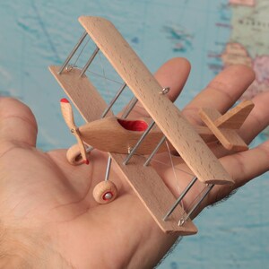 May include: A miniature biplane model crafted from light-colored wood, featuring two stacked wings, a small propeller with red tips, and thin metal struts. The cockpit interior is lined with red fabric, and the landing gear includes two small wheels.