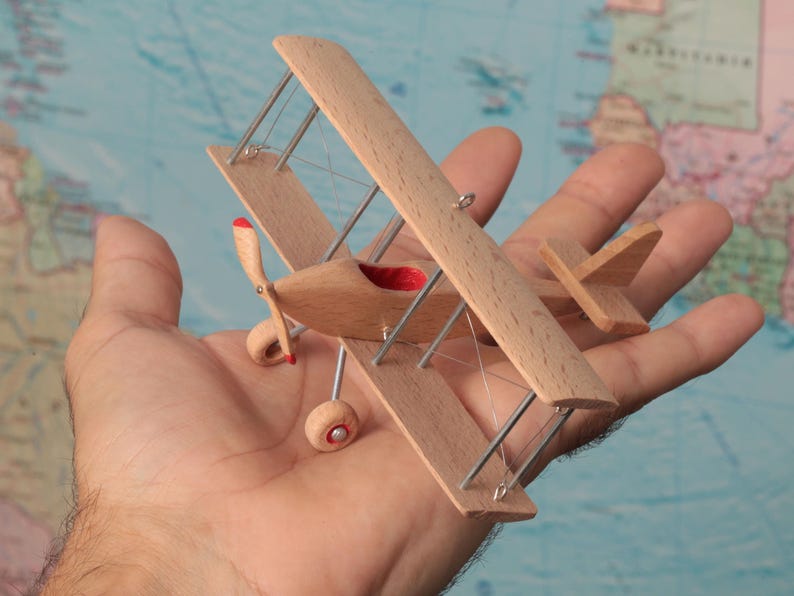 May include: A miniature biplane model crafted from light-colored wood, featuring two stacked wings, a small propeller with red tips, and thin metal struts. The cockpit interior is lined with red fabric, and the landing gear includes two small wheels.