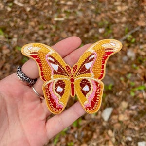 May include: Embroidered butterfly patch with a detailed design. The patch features a yellow background with red, brown, pink, and white accents. The butterfly's wings are spread, showcasing intricate patterns and textures. The patch is likely for clothing or accessories.
