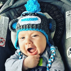 May include: A baby wearing a gray and blue crocheted monkey hat with a blue pom-pom on top. The hat has white accents and long blue and white braided ties.