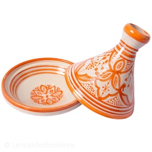 May include: A white ceramic tagine with an orange glaze and intricate floral designs. The tagine has a round base and a conical lid with a small knob on top.