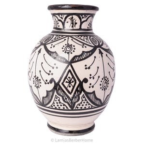 Può includere: Vaso in ceramica bianca con motivi geometrici e floreali neri. Il vaso ha un corpo rotondo e un collo stretto.
