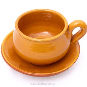 Może przedstawiać: Ręcznie wykonany zestaw filiżanki i spodka ceramicznego. Filiżanka ma zaokrąglony kształt z uchwytem, a spodek jest okrągły. Oba elementy są glazurowane w ciepłym, ziemistym pomarańczowym kolorze. Zestaw przeznaczony do herbaty lub kawy.