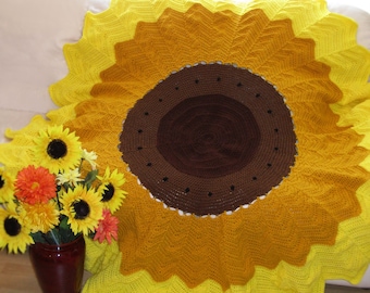 May include: A crocheted sunflower blanket with yellow, brown, and white yarn. The blanket is in the shape of a sunflower with a brown center and yellow petals.
