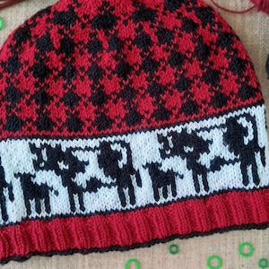 Puede incluir: Un gorro de punto rojo y negro con una banda blanca que presenta un patrón repetido de renos estilizados. El gorro está hecho con un hilo grueso y tiene una textura suave y acogedora.