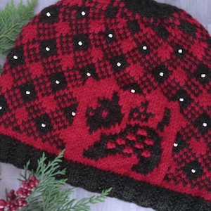 May include: A red and black knitted hat with a repeating diamond pattern and white dots. The hat features a black silhouette of a dog on the front. The brim is black. This winter hat is perfect for cold weather.