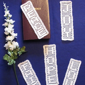 May include: Five white crocheted bookmarks on a blue surface. The bookmarks are rectangular and have a delicate, lacy pattern. One bookmark is on top of a closed brown book with the word "BIBLE" printed on the cover.
