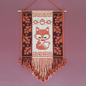Puede incluir: Un tapiz de pared tejido con un diseño de zorro. El tapiz es principalmente naranja, marrón y blanco, con un patrón geométrico repetido en los lados y una franja en la parte inferior. El zorro está centrado.