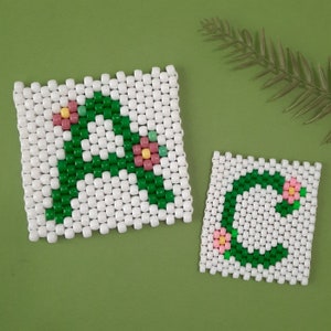 May include: Two white beaded coasters with green letters and pink flowers. The coasters spell out the letters "A" and "C".