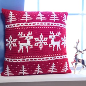 May include: A red crocheted pillow with white Christmas tree and reindeer patterns. The pillow is square-shaped and sits on a white surface. The design is a festive holiday theme.