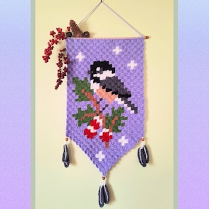 Könnte beinhalten: Ein lilafarbener gehäkelter Wandbehang mit einem Vogel- und Beerenmotiv. Der Vogel ist ein Meisen mit schwarzen, weißen und grauen Federn. Die Beeren sind rot und grün. Der Behang hat unten Quasten.