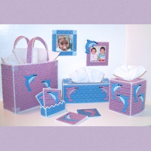 May include: A set of pink and blue tissue box covers and coasters with a dolphin design. The tissue box covers have a white tissue inside. The coasters are in a holder. There are also two framed pictures with photos of children.
