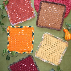 May include: Five crocheted squares in different colours, including orange, brown, yellow, red, and burgundy. Each square has a white crocheted design in the centre.