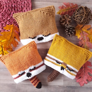Puede incluir: Tres gorros de panda rojo de punto en diferentes tonos de naranja y amarillo. Cada gorro tiene un borde blanco y ojos negros. Los gorros están sobre una superficie de madera con hojas de otoño y piñas.