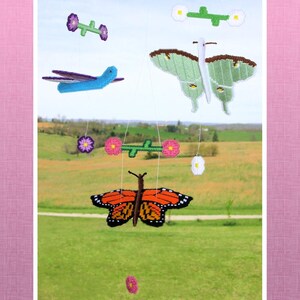 May include: A handmade mobile featuring various colorful, stitched insect and floral designs. The mobile includes a butterfly with orange and black wings, a green butterfly, a blue dragonfly, and several flower accents. The background shows a green field and a blue sky.