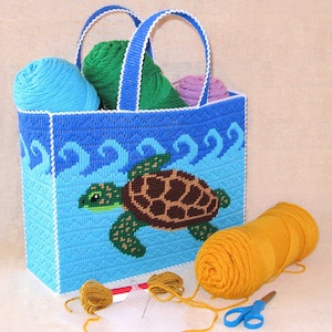 May include: A blue canvas tote bag with a white trim and a sea turtle design. The bag is filled with yarn balls in various colors, including blue, green, and purple. There is also a ball of yellow yarn and a pair of scissors on the table.