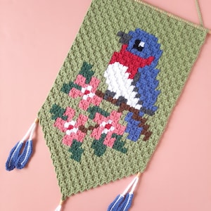 May include: A crocheted wall hanging featuring a bluebird with red and white accents perched on a branch with pink flowers. The hanging has green tassels.