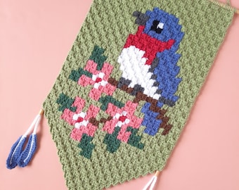 May include: A crocheted wall hanging featuring a bluebird with red and white accents perched on a branch with pink flowers. The hanging has green tassels.