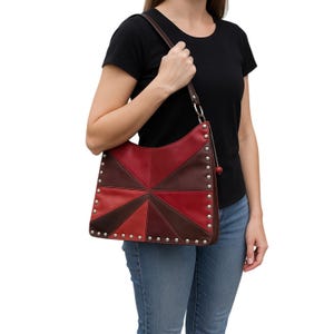 May include: A red, brown, and burgundy leather shoulder bag with a geometric design. The bag features silver-colored studs along the edges and a brown leather strap. The bag is held by a person wearing a black t-shirt and blue jeans.