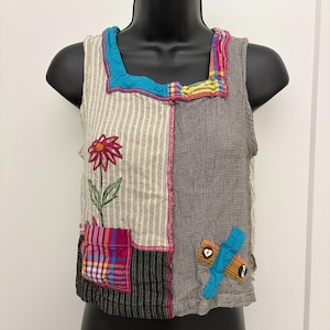 Könnte beinhalten: Ein ärmelloses Oberteil im Patchwork-Design mit quadratischem Ausschnitt. Die linke Seite hat cremefarbene und schwarze vertikale Streifen mit einer roten Blumenstickerei und einer karierten Tasche. Die rechte Seite ist grau mit einer Applikation.