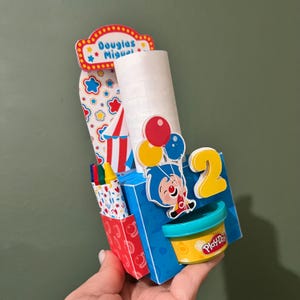 Puede incluir: Una caja de cartón azul y roja con un personaje de payaso que sostiene globos. La caja tiene un contenedor de Play-Doh, crayones y un rollo de papel. La caja está decorada con estrellas, una tienda de campaña y el número 2. La caja dice "Douglas Miguel" en la parte superior.
