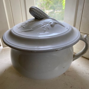 May include: White ceramic chamber pot with a lid. The lid has a raised design of a corn cob and leaves. The pot has a single handle.