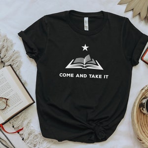 May include: A black short-sleeved t-shirt featuring a white graphic of an open book with a single star positioned above it. Below the book, the text reads "COME AND TAKE IT" in white capital letters. The shirt has a crew neckline and short sleeves.