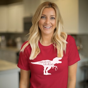 May include: A red t-shirt with a white graphic of a Tyrannosaurus Rex holding a book, with the text "Librarian Saurus". The shirt is made of a soft material.