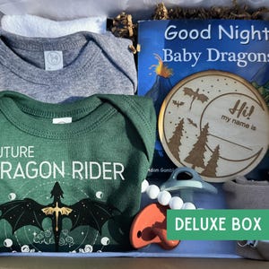 May include: A gift box containing a grey baby bodysuit, a green baby bodysuit with a dragon graphic and the text Future Dragon Rider, a Good Night Baby Dragons book, a wooden name plaque, two pacifiers, and a beige fabric item with a metal clip.