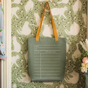 a green tote bag hanging on a wall with a pattern of white cats. The bag has a list of information printed on it, including the title, author, title, and date due. The wall behind the bag has a floral pattern.
