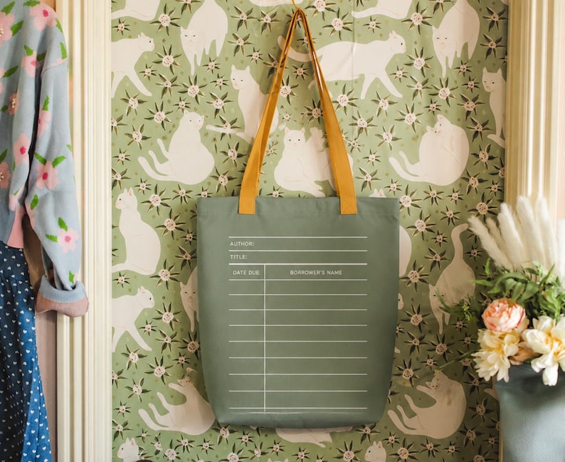 a green tote bag hanging on a wall with a pattern of white cats. The bag has a list of information printed on it, including the title, author, title, and date due. The wall behind the bag has a floral pattern.