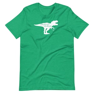 May include: A green t-shirt featuring a white graphic of a Tyrannosaurus Rex holding a book. The text "Librarian Saurus" is printed above the dinosaur. The shirt is made of a soft material.