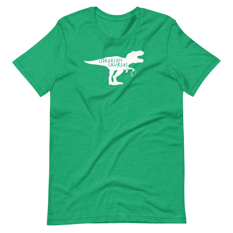 May include: A green t-shirt featuring a white graphic of a Tyrannosaurus Rex holding a book. The text "Librarian Saurus" is printed above the dinosaur. The shirt is made of a soft material.