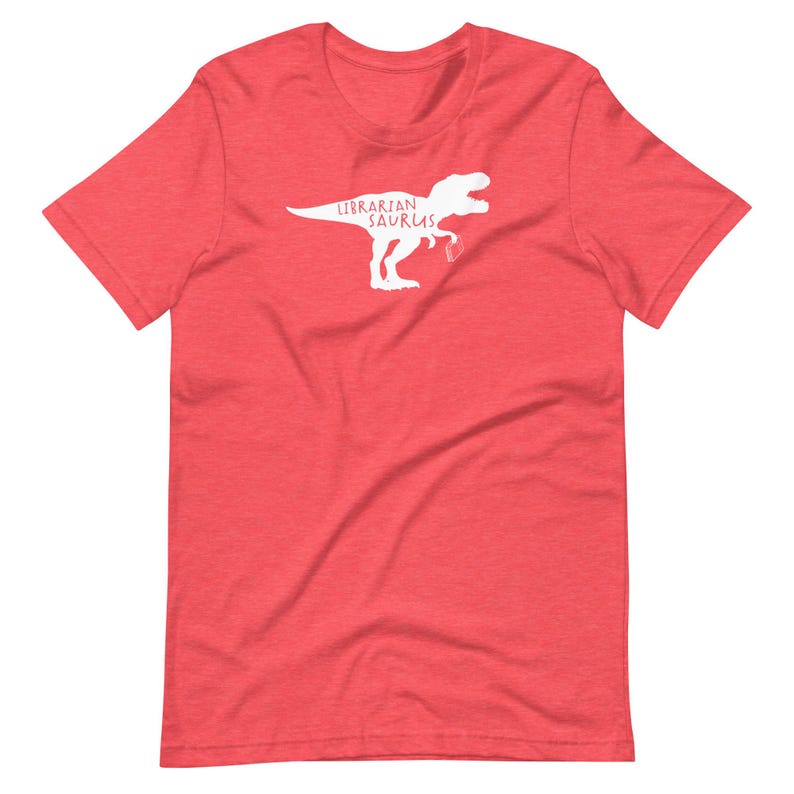 May include: Red t-shirt with a white graphic of a Tyrannosaurus Rex holding a book and the words "Librarian Saurus". The shirt is made of a soft material and has short sleeves and a crew neck.