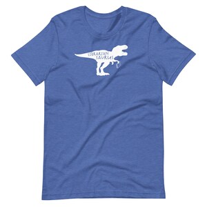 May include: Blue t-shirt with a white graphic of a Tyrannosaurus Rex holding a book. The text "Librarian Saurus" is printed above the dinosaur. The shirt is made of a soft material.