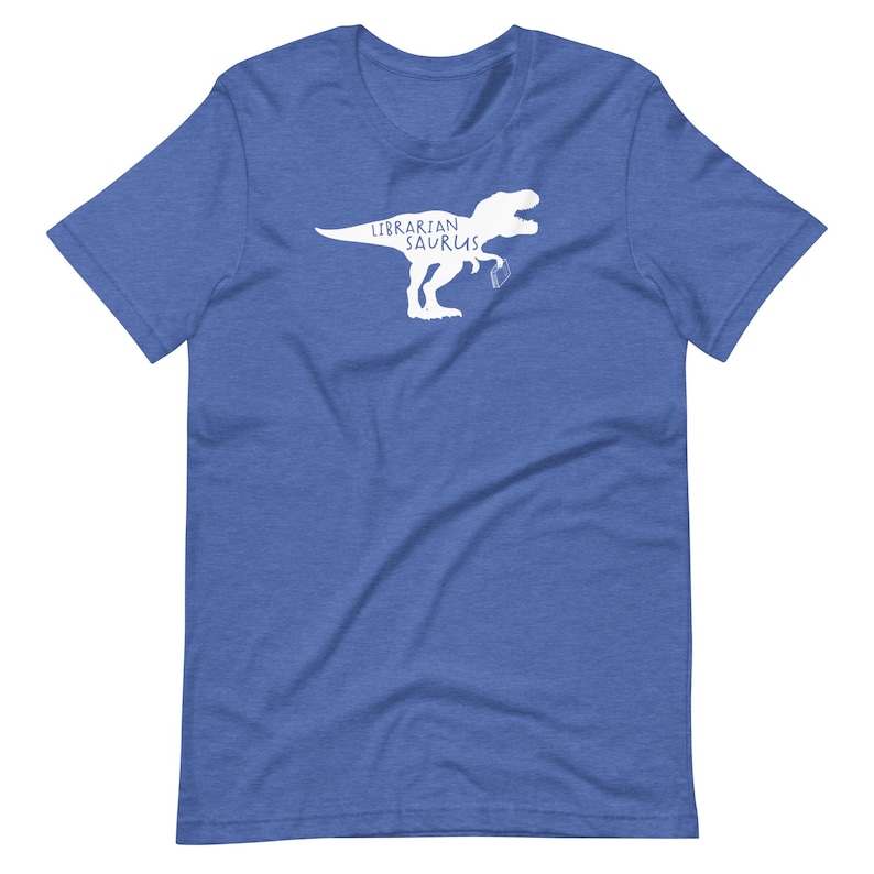 May include: Blue t-shirt with a white graphic of a Tyrannosaurus Rex holding a book. The text "Librarian Saurus" is printed above the dinosaur. The shirt is made of a soft material.