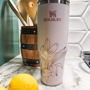 Puede incluir: Un termo de viaje Stanley rosa claro con un dise&ntilde;o floral y el logotipo de Stanley. El termo tiene una tapa rosa claro y un cierre plateado. Dos limones amarillos y cucharas de madera est&aacute;n en el fondo.