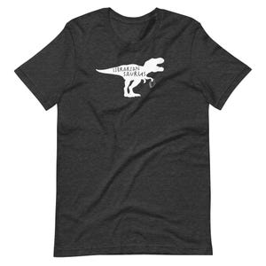 May include: Dark gray t-shirt featuring a white graphic of a Tyrannosaurus Rex holding a book and the words "Librarian Saurus". The shirt is made of a soft material and has short sleeves and a crew neck.