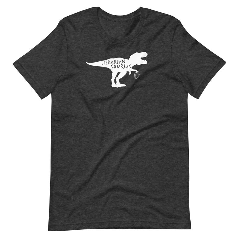 May include: Dark gray t-shirt featuring a white graphic of a Tyrannosaurus Rex holding a book and the words "Librarian Saurus". The shirt is made of a soft material and has short sleeves and a crew neck.
