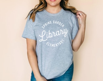 May include: A light grey short-sleeved t-shirt featuring white screen-printed text on the chest that reads Spring Garden Library Elementary in a circular design. The shirt has a crew neckline and a standard fit, made of a heathered fabric.